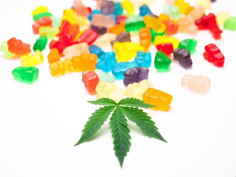 Vibrant gummy bears scattered alongside a cannabis leaf on a white background.
